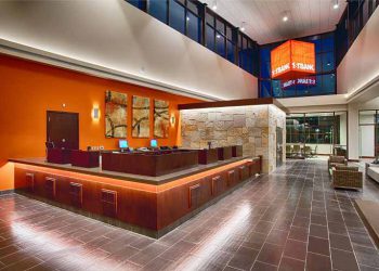 Open plan with with orange wall behind large corner wood work station for bank tellers.